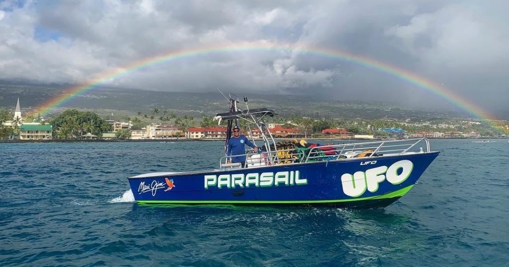 Blue Speed Boat with UFO Parasail written on the side in the ocean with Kailua Kona town and a rainbow in the background